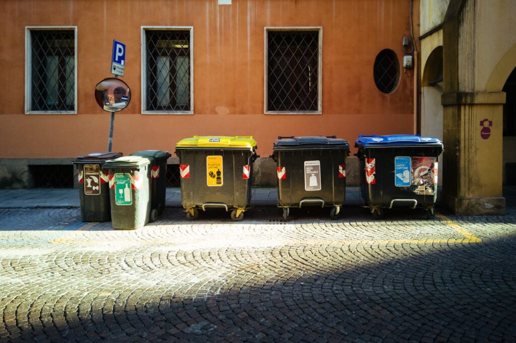 Five recycling bins on a sunny cobblestone street against an orange building.